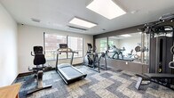 Located at 2732 Forest Ave in Kansas City, MO, this fitness center offers a focused and well-equipped exercise space designed to support a well-rounded workout routine. The room features a patterned carpet tile floor in muted blue and grey tones, lending a modern aesthetic throughout. Large windows along one wall flood the space with natural light and provide views of the surrounding neighborhood, creating an open and energizing atmosphere.

The exercise room is outfitted with a variety of cardio equipment, including a treadmill, an elliptical trainer, and a recumbent cycle, allowing for diverse cardiovascular training options. A multi-function cable weight machine with an adjustable bench provides strength training capabilities. A full wall-length mirror enhances the sense of space and allows users to monitor their form during workouts.

A wooden bench and wall-mounted coat hooks offer a functional area for personal belongings, while framed wall art adds a refined touch to the environment. Flush ceiling light panels ensure consistent, even illumination throughout the room. A glass-paneled door connects the fitness center to an adjacent corridor, maintaining an open and accessible feel. The space is neatly designed to maximize utility within its compact footprint, making it a practical and comfortable fitness amenity.