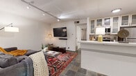 This single-floor unit offers a thoughtfully arranged layout comprising a living room, kitchen, and bedroom across a compact but functional footprint. Dark slate tile flooring runs consistently throughout the space, lending a cohesive and modern aesthetic.

The living room serves as the main gathering area, featuring a window dressed with floor-length gray curtains that allow natural light to filter in. Track lighting on the ceiling provides adjustable illumination, complementing the room's open feel. The living area connects directly to the kitchen via an open-concept design, separated by a peninsula counter with a light stone surface that doubles as a breakfast bar.

The kitchen is equipped with stainless steel appliances, including a refrigerator, range, and dishwasher. White cabinetry with glass-front upper panels provides ample storage, while a tile backsplash adds a subtle design detail. The light stone countertops extend generously along the galley-style workspace.

The bedroom offers a comfortable retreat with a ceiling fan, floor-length curtains, and a walk-in closet with built-in shelving. Upholstered wall panels behind the bed area serve as a distinctive architectural accent, adding depth and texture to the room.