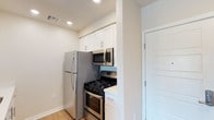 This single-story space includes a thoughtfully designed kitchen that maximizes functionality within a compact footprint. The kitchen is outfitted with a full suite of stainless steel appliances, including a full-size refrigerator, a gas range with oven, and an over-the-range microwave, offering a complete and modern cooking setup. White shaker-style upper cabinets with brushed nickel hardware provide clean, classic storage above the appliance alcove, contributing to a bright and tidy aesthetic. Light-toned hardwood-style flooring runs throughout, adding warmth to the neutral color palette of white walls and soft recessed lighting overhead. The recessed ceiling lights ensure the space feels well-lit and inviting. A white paneled door adjacent to the kitchen leads to an additional room or closet, suggesting practical storage or utility access nearby. The overall design is clean, minimal, and functional, with a cohesive use of light tones and modern finishes that create a fresh, welcoming atmosphere suited to everyday living.