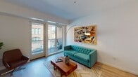 This well-designed single-floor residence offers a thoughtful layout that balances open communal spaces with comfortable private retreats. The open-concept living area is anchored by light hardwood flooring and features a tufted teal sofa, a live-edge wood coffee table, and bold abstract wall art. Floor-to-ceiling glass doors flood the space with natural light and open directly onto a private balcony, offering views of the surrounding urban landscape.

The kitchen is a standout feature, showcasing sleek white cabinetry, quartz countertops, stainless steel appliances, and a large kitchen island with bar seating — all illuminated by recessed lighting and a striking pendant fixture. The space is both functional and stylish, ideal for cooking and entertaining.

The primary bedroom is a serene retreat, distinguished by a rich forest-green accent wall, a plush upholstered headboard, and a walk-in closet. A second bedroom offers a bright and airy atmosphere, with large windows framing urban views and a warm mid-century dresser. The full bathroom is finished in crisp white, featuring a wide vanity with ample storage, a large mirror, and a shower. An in-unit washer and dryer add everyday convenience to this polished, modern residence.