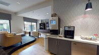 This well-appointed single-floor lounge space combines contemporary design with practical amenities, creating a welcoming environment for guests or residents. At the heart of the space is a sleek beverage station, prominently featuring a Starbucks coffee machine alongside an AquaMaestro water dispenser, set against a striking geometric-textured accent wall in crisp white. The counter provides ample workspace with dark cabinetry below for storage.

The lounge area is divided into two distinct seating zones. One section features a relaxed arrangement of cream-upholstered armchairs paired with a glass-top coffee table, all set on warm hardwood-style flooring. This area benefits from abundant natural light streaming through large sliding glass doors that open directly onto a private outdoor patio, complete with patio furniture and lush planters. The second seating zone offers a more social setting, with tufted leather chairs in warm amber tones, a striped sofa accented with blue throw pillows, and abstract artwork adorning the walls.

A built-in dark espresso wood media unit dominates one wall, housing a large flat-screen television, display shelving with decorative accents, and storage drawers below. Additional wall-mounted screens are positioned throughout the space. Recessed lighting throughout provides a clean, polished finish, and the overall aesthetic balances modern sophistication with a comfortable, inviting ambiance.