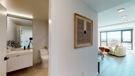 This single-floor studio is thoughtfully designed with a modern aesthetic, blending clean lines and functional living spaces. The open-plan layout seamlessly connects the living, sleeping, and kitchen areas, creating an efficient and cohesive environment. Large windows flood the space with natural light while offering sweeping city views, adding a sense of openness and depth.

The kitchen is a standout feature, showcasing dark lower cabinetry paired with crisp white upper cabinets, a white tile backsplash, and bright countertops. A stainless steel sink with matte black fixtures and a full suite of appliances, including an electric range and built-in microwave, complete this well-appointed culinary space. Light wood-tone flooring runs throughout, tying the kitchen to the rest of the unit.

The living and sleeping area benefits from an exposed concrete structural column, lending an industrial character to the otherwise minimalist design. A large window frames panoramic city views, enhancing the airy ambiance.

The bathroom is finished with large-format beige tiles, a floating white vanity with a vessel sink, matte black hardware, and a full-length mirror that amplifies the sense of space. A sliding door conceals a stacked in-unit washer and dryer, offering practical convenience without disrupting the clean aesthetic of the living area.