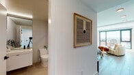 This single-floor studio is thoughtfully designed with a modern aesthetic, blending clean lines and functional living spaces. The open-plan layout seamlessly connects the living, sleeping, and kitchen areas, creating an efficient and cohesive environment. Large windows flood the space with natural light while offering sweeping city views, adding a sense of openness and depth.

The kitchen is a standout feature, showcasing dark lower cabinetry paired with crisp white upper cabinets, a white tile backsplash, and bright countertops. A stainless steel sink with matte black fixtures and a full suite of appliances, including an electric range and built-in microwave, complete this well-appointed culinary space. Light wood-tone flooring runs throughout, tying the kitchen to the rest of the unit.

The living and sleeping area benefits from an exposed concrete structural column, lending an industrial character to the otherwise minimalist design. A large window frames panoramic city views, enhancing the airy ambiance.

The bathroom is finished with large-format beige tiles, a floating white vanity with a vessel sink, matte black hardware, and a full-length mirror that amplifies the sense of space. A sliding door conceals a stacked in-unit washer and dryer, offering practical convenience without disrupting the clean aesthetic of the living area.