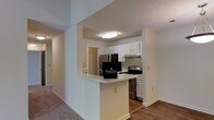This single-floor residence offers a thoughtfully designed layout that balances functionality and comfort. The kitchen serves as a central highlight, outfitted with stainless steel appliances including a refrigerator, range, and dishwasher, complemented by crisp white cabinetry that provides ample storage. A peninsula breakfast bar separates the kitchen from the adjoining living space, creating a natural flow between areas while offering a casual spot for dining or entertaining. Light-toned countertops contrast neatly with the hardwood-style flooring that runs through the kitchen and connecting areas, while plush carpeting adds warmth to the living spaces. The walls are finished in a clean, neutral palette that enhances the sense of brightness throughout. Flush-mount ceiling lighting keeps the kitchen well-illuminated, and a range hood above the stove adds a practical touch. An open corridor connects the main living areas, contributing to an airy, cohesive feel across the floor plan. Overall, the space presents a clean, move-in-ready environment with classic finishes and a layout well-suited for everyday living.