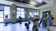 This fitness center is a thoughtfully designed, single-floor facility built to support a comprehensive workout experience. The space features a tray ceiling with a bold dark upper section accented by recessed and track lighting, creating a modern and energizing atmosphere. Large, multi-pane windows line the walls, flooding the interior with natural light and offering pleasant views of surrounding greenery, enhancing the overall workout environment. The flooring is a smooth, light-toned surface well-suited for fitness use. The facility is outfitted with a range of equipment, including a cable and pulley strength machine in the foreground, a stationary recumbent bike, and additional cardio and strength machines positioned throughout the room. Mirrors are strategically placed along the walls to maximize the sense of space and allow users to monitor their form. A framed notice or rules placard is mounted near the entrance, reflecting an organized and well-managed facility. The overall ambiance is clean, functional, and professional, making this fitness center a practical and inviting space for a full range of exercise routines.