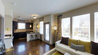 Located at 11775 Wadsworth Boulevard in Broomfield, Colorado, this single-floor residence offers a thoughtfully designed open-concept layout that seamlessly connects the living and kitchen areas. Rich, dark hardwood flooring runs throughout the space, lending warmth and continuity to the interior.

The kitchen is outfitted with sleek dark cabinetry, a neutral countertop, and a peninsula with bar-height seating — ideal for casual dining or entertaining. Track lighting and a pendant light above the peninsula provide focused illumination, while a window above the sink invites natural light into the cooking space.

The living area is generous and light-filled, benefiting from multiple large windows dressed with patterned drapes and horizontal blinds that offer both style and privacy. The abundance of natural light creates an airy, welcoming ambiance. A floor lamp adds a warm accent in the corner, complementing the neutral and earth-toned décor.

The overall aesthetic is modern and cohesive, with a neutral color palette accented by yellow and brown tones that add character without overwhelming the space. The open flow between functional zones makes this residence feel expansive and versatile, suited to a variety of lifestyles.