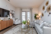 This thoughtfully designed single-floor residence offers a cohesive blend of modern aesthetics and functional living spaces. The open-concept living area serves as the heart of the home, featuring warm hardwood flooring, neutral-toned walls, and an eye-catching display of gold and silver starburst wall art that adds a bold, artistic focal point above a comfortable sofa. Large French doors flood the space with natural light and provide access to a private balcony, enhancing the airy, welcoming ambiance. A wall-mounted flat-screen television sits atop a sleek walnut media console, completing the living area's contemporary look. The kitchen is outfitted with rich, dark wood cabinetry, granite countertops, a white subway tile backsplash, and a full suite of stainless steel appliances, including a refrigerator, electric range, and built-in microwave — combining both style and practicality. The bedroom is a calming retreat, anchored by a soft teal accent wall adorned with three decorative sunburst mirrors. Mid-century modern nightstands flank the bed on either side, contributing to the room's relaxed yet refined character. The bathroom maintains a clean, minimal aesthetic with white tile, a shower-tub combination, and neutral wall tones that create a calm and functional space.