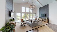 This single-story clubhouse is designed to impress, combining modern farmhouse and contemporary aesthetic elements throughout its well-appointed interior. The centerpiece of the space is a dramatic great room with soaring vaulted ceilings accented by exposed beams and floor-to-ceiling windows that frame views of the outdoor pool and surrounding landscape. A striking black iron chandelier anchors the room above a generous seating area, while a charcoal shiplap feature wall houses a mounted television and rustic media console. Light hardwood flooring runs throughout, lending warmth and continuity across zones.

Transitioning deeper into the clubhouse, a rope bead curtain partition elegantly defines a dining area furnished with a wooden table and mixed-seating chairs, maintaining visual openness while creating distinct functional spaces. Leathered concrete-finish desks and upholstered chairs outfit a dedicated workspace area, its teal accent wall adorned with bold, eclectic artwork. A kitchenette is accessible from the main area, offering added convenience. Throughout, the interplay of natural light, curated art, and thoughtful architectural details creates a welcoming, vibrant community hub.