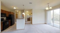 This single-floor unit offers a welcoming open-concept layout that seamlessly connects the kitchen and living areas, creating a spacious and functional environment. The kitchen is highlighted by a white peninsula with a sleek countertop, pendant lighting overhead, and warm wood-toned cabinetry. A full-size refrigerator anchors the space, while tile flooring transitions smoothly into plush carpeting in the living area. The open living space is generously sized, bathed in ambient light from ceiling fixtures, and features neutral-toned walls that lend a clean, versatile aesthetic. A hallway visible from the main living area leads to additional rooms, including what appears to be a bathroom with dark wood vanity cabinetry, adding a warm, classic touch. The unit benefits from ceiling vents throughout, ensuring comfortable climate control. One of the standout features of this space is its outlook — windows frame a picturesque view of mature deciduous trees displaying vibrant autumn foliage, alongside an open natural landscape and a well-maintained parking area below. This natural backdrop provides a serene, park-like setting that enhances the overall ambiance of the unit, making it feel connected to the outdoors despite being an enclosed residential space.
