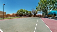 Located at 9300 Montana Ave in El Paso, Texas, this outdoor recreational facility centers around a well-maintained, full-size tennis court finished in a classic green and red hard-court surface with clearly marked white boundary lines and a regulation net. The court is enclosed by chain-link fencing that defines the playing area while maintaining open sightlines to the surrounding landscape. Mature trees line the perimeter, offering natural shade and a lush, verdant backdrop against El Paso's characteristically clear blue skies. A standout feature of the facility is the pair of teal shade canopy structures positioned courtside, providing a comfortable shaded seating and gathering area for players and spectators alike. Raised wooden planters with desert-adapted landscaping add aesthetic detail to the courtside rest area. Lighting poles are present, suggesting the court may be usable during evening hours. The surrounding grounds include well-kept green lawn areas and desert-friendly plantings, complementing the warm earth-toned adobe-style residential buildings visible in the background. The overall ambiance is inviting and well-suited for recreational play, combining practical amenities with a pleasant outdoor environment reflective of the sunny Southwest setting.