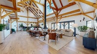 The Barn is a thoughtfully designed two-story space that masterfully balances rustic heritage with contemporary functionality. The defining architectural feature is the exposed timber post-and-beam framework, which runs throughout both floors, lending warmth and structural drama to every area.

The main level opens into a generous great room anchored by a suspended central fireplace and flanked by multiple seating arrangements on wide-plank hardwood floors. Large glass doors along the perimeter invite abundant natural light and provide views to outdoor areas. A long communal dining table anchors an adjacent gathering zone, framed by the same signature timber trusses overhead.

Private rooms accessible via sleek black sliding barn doors offer quieter settings, including a compact workspace with carpeted flooring and sage-green walls. A streamlined kitchenette features light countertops, matte cabinetry, open wood shelving, and modern appliances. Curated artwork is displayed throughout, adding visual interest.

The second floor, reached via a staircase with modern metal railings, mirrors the timber-framed aesthetic and overlooks the main level, reinforcing the open, interconnected nature of the space.