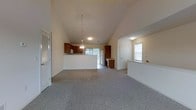 This single-floor home offers an open-concept design that seamlessly connects the living area and kitchen, creating a spacious and welcoming environment. The soaring vaulted ceilings lend a generous sense of height and airiness throughout the main living space, which is finished with soft, wall-to-wall carpeting underfoot.

The kitchen is well-defined by its warm, rich wood cabinetry that provides ample storage, complemented by a breakfast bar or pass-through counter that reinforces the open flow between the kitchen and living area. A pendant light fixture hangs above, adding a functional yet stylish focal point. Natural light filters in through windows, keeping the space bright and inviting.

A dedicated entry area with a paneled door provides a clear point of arrival, while an additional window nearby draws in further natural light. The neutral color palette throughout — from the walls to the flooring — creates a clean, versatile backdrop well-suited to a variety of furnishing styles.

Overall, this home balances practicality with comfort, offering an open, light-filled layout anchored by standout architectural details like the vaulted ceiling and thoughtfully connected living and kitchen spaces.