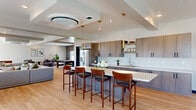 This expansive single-floor community space offers a thoughtfully designed open-plan layout that seamlessly integrates multiple functional zones beneath coffered ceilings with recessed lighting and ceiling-mounted HVAC units. Light hardwood flooring flows throughout, unifying the space and enhancing its bright, contemporary aesthetic.

The fully equipped kitchen serves as a central anchor, featuring flat-front cabinetry in a warm wood-grain finish, white quartz countertops, a stainless steel French door refrigerator, and a generous island with bar seating for several guests. Gold-accented pendant lights add an elevated touch above the island.

Adjacent to the kitchen, a dining area with long communal tables and modern black bistro chairs provides an ideal setting for group gatherings. The lounge area offers generous sectional seating, a wall-mounted flat-screen TV, and large windows that invite abundant natural light and exterior views. A foosball table and game area cater to recreational use.

Separate intimate seating nooks are thoughtfully arranged throughout, including a cozy lounge zone with rounded swivel chairs and a formal sitting area furnished with plush accent chairs, a patterned area rug, and a linear gold chandelier. Curated greenery and artwork complete the polished, welcoming ambiance.