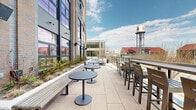 Located on the fourth floor of a modern Baltimore building, this expansive outdoor terrace offers a well-appointed open-air amenity space with striking urban views. The terrace is finished with large-format neutral tiles underfoot, creating a clean, contemporary aesthetic throughout.

The space is thoughtfully organized into distinct seating zones. A lounge area features a generous sectional sofa in light gray upholstery paired with a slatted coffee table, offering a relaxed setting with panoramic views of the surrounding Baltimore streetscape, including a distinctive landmark water tower. Bistro-style pedestal tables with casual bar seating and tall chairs provide additional gathering spots suited for individual or small-group use.

Integrated planter beds line portions of the terrace, filled with ornamental grasses and seasonal plantings that add a natural, softening element to the urban environment. A sleek metal railing runs along the perimeter, ensuring unobstructed sightlines across the cityscape while maintaining an open, airy feel.

The terrace is flanked by the building's brick and glass façade, which features large industrial-style glazed garage doors that connect the outdoor space to the interior. Outdoor speakers and wall-mounted lighting are also present, supporting both daytime and evening use of this versatile outdoor space.