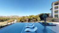 This expansive outdoor amenity area centers around a stunning infinity-edge pool designed to seamlessly blend with the natural landscape beyond. The pool's vanishing edge faces an unobstructed panorama of rolling, tree-covered hills and distant low-rise buildings, creating a striking visual connection between the water and the horizon. Three sculptural, wave-form in-water lounge chairs are positioned along the infinity edge, allowing occupants to recline directly in the shallow water while taking in the views.

The pool deck is finished in smooth, light-toned concrete and is generously lined with white slatted chaise lounges positioned beneath a covered overhang supported by large cylindrical columns. The overhang features a warm wood-paneled ceiling, providing shaded relief directly adjacent to the pool's edge. A separate spa with water features and an in-pool island planter with native yucca and ornamental grasses add visual interest and a resort-like character to the space.

The pool area is bordered by sleek black metal fencing that maintains sightlines to the surrounding landscape. The adjacent multi-story building showcases contemporary architecture with large windows, mixed exterior materials, and private balconies that overlook the amenity deck. The overall atmosphere is polished, open, and oriented toward outdoor living.