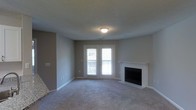 This single-floor residential space offers a welcoming and functional layout designed for comfortable everyday living. The living area serves as the heart of the home, finished with wall-to-wall carpeting in a neutral tone that adds warmth underfoot. Soft gray walls create a calm, cohesive backdrop that complements the clean white trim and baseboards throughout. A fireplace with a classic white mantel anchors one end of the room, providing a natural focal point and a cozy atmosphere. Opposite the fireplace, a set of French doors floods the space with natural light, offering a bright and airy feel while providing access to an outdoor area. The doors are dressed with horizontal blinds, allowing for flexible light control and privacy. Ceiling lighting ensures the room remains well-lit regardless of the time of day. The open feel of the living area is further enhanced by a glimpse of additional counter space — likely a kitchen transition — visible along one edge of the room, suggesting a practical and connected floor plan. Overall, this space presents a clean, move-in-ready environment with a neutral palette and key features that balance comfort and style.