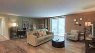 This single-floor space centers around a welcoming living area finished with warm-toned hardwood flooring that runs throughout. The room is anchored by a comfortable tan sofa accented with decorative pillows in earthy orange and brown hues, paired with a round dark ottoman serving as a coffee table. A sliding glass door floods the space with natural light and offers a lush, green outdoor view, creating a bright and airy atmosphere. A patterned accent chair is positioned near the door, adding a secondary seating option. The walls are painted in warm, muted earth tones — a combination of sage green and terracotta — contributing to a cozy, grounded ambiance. A wall-mounted air conditioning unit provides climate control. Along one wall, a cork board and desk area suggest a functional workspace integrated into the living space. A tall mirror leans against the wall, visually expanding the room. The overall design balances practicality with comfort, making it a versatile, inviting environment suited for everyday living and relaxing.