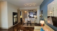This single-floor Dallas residence showcases a thoughtfully designed open-concept layout that connects the living, dining, and kitchen areas into one cohesive space. Dark hardwood flooring runs throughout, anchoring the interior with warmth and contrast against the light-colored walls and white kitchen surfaces.

The kitchen is appointed with dark cabinetry, granite countertops, stainless steel appliances including a French door refrigerator, and pendant lighting that hangs above the peninsula-style counter. The peninsula provides additional seating and prep space while maintaining visual openness between the kitchen and the rest of the living area.

Adjacent to the kitchen, a compact dining area accommodates a round table with upholstered chairs, set atop a patterned area rug. Track lighting above enhances the functional brightness of this zone. A bold accent wall in muted blue adds a contemporary design element to the space.

The living area extends naturally from the dining space, offering a relaxed, connected feel throughout. A large floor mirror near the entryway amplifies natural light and adds visual depth. The overall ambiance is modern and polished, with a clean, urban aesthetic well-suited to Dallas city living.