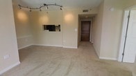 This single-floor unit features an open-concept layout that integrates the living area and kitchen into a cohesive, functional space. The living room is finished with wall-to-wall carpet in a neutral beige tone, providing a soft foundation that complements the warm, cream-colored walls throughout. A curved track lighting fixture overhead illuminates the living space, offering directed lighting that enhances the room's ambiance.

A pass-through window connects the living area to the kitchen, offering a glimpse of the appliances, including a black refrigerator and cabinetry with a light countertop. This design element maintains a sense of openness while defining the separate cooking space.

A hallway leads from the main living area to the front entry, where a solid dark wood door serves as a striking focal point. The corridor is finished with the same neutral carpet and walls, maintaining a consistent aesthetic throughout. Ceiling vents are visible, indicating a central HVAC system. The overall design is clean, simple, and move-in ready, suited for those seeking a straightforward, low-maintenance living environment.