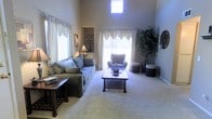 This single-story residence is defined by its open-concept design, where the living, dining, and kitchen areas flow together to create a cohesive and functional living environment. The living room immediately draws attention with its dramatic vaulted ceilings, which lend a sense of spaciousness and grandeur to the space. Natural light pours in through a combination of large sliding glass doors, standard windows, and a strategically placed clerestory window positioned high on the wall, ensuring the room remains bright throughout the day. Neutral-toned carpet covers the living area floor, contributing to a warm and inviting ambiance, while the dining area transitions to hard surface flooring.

The kitchen is positioned adjacent to the dining area and features a peninsula with a dark granite countertop, providing a natural visual separation between cooking and living spaces. Light-toned cabinetry and a built-in refrigerator contribute to a clean, organized aesthetic. A ceiling fan is installed in the living area, offering both comfort and a functional design element. The overall color palette throughout the home consists of warm beige and neutral tones, creating a calm, cohesive atmosphere across all areas.