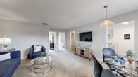 This single-floor residence offers a thoughtfully organized layout with clearly defined living spaces. The living room serves as the central hub of the home, featuring soft wall-to-wall carpeting, neutral gray walls, and a textured ceiling. The open arrangement comfortably accommodates both a seating area and a dining space, with a wall-mounted television and pendant lighting adding functional detail. Natural light enters through multiple openings, and direct sightlines connect the living area to the bedrooms beyond.

The galley-style kitchen is efficiently designed with gray cabinetry lining both walls, white countertops, a double-basin sink, and light wood-look flooring. White appliances including a refrigerator and range are integrated seamlessly into the layout, while a window provides additional brightness.

Two bedrooms are accessible from the main living area. Each room is carpeted, finished in soft neutral tones, and features sliding glass doors with vertical blinds that allow natural light and outdoor views. The rooms offer generous space for full-sized furniture.

The bathroom includes wood-look flooring, a white vanity with a framed mirror, and a tub-shower combination accented by a bold striped curtain. The overall ambiance is clean, functional, and move-in ready.