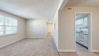 Located at 705 S Beach St in Daytona Beach, Florida, this single-level one-bedroom, one-bathroom unit offers a functional and efficient layout. The living room serves as the heart of the space, featuring neutral-toned carpet, a textured ceiling, and a pass-through window detail on one wall, with a hallway leading toward the entry door and additional rooms. Natural light filters in through a window fitted with blinds, giving the living area a bright and open feel. The flooring transitions from carpet in the main living area to hard surface flooring near the entryway and kitchen zone, as visible in the doll-house view. The bedroom is a comfortable, well-proportioned room with plush carpeting, a ceiling fan with light fixture, a window allowing natural light, and access to a dedicated closet. The bathroom is clean and straightforward, featuring a full soaking tub with a classic white tiled surround and chrome fixtures. Throughout the unit, neutral walls, white trim, and textured ceilings maintain a cohesive, understated aesthetic. The hallway connects the main living spaces efficiently, completing a practical and livable floor plan well-suited for everyday comfort in the heart of Daytona Beach.