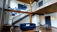 This impressive two-story space immediately captures attention with its soaring double-height ceilings and rich, exposed wooden beams that lend a warm, industrial character to the interior. The main level centers around an inviting living area furnished with a deep navy velvet sofa and complementary seating, anchored by hardwood flooring and a cowhide accent rug. A bold sputnik-style chandelier with globe bulbs serves as a dramatic focal point, suspended elegantly between the two floors. A sleek metal-railed staircase with clean, modern lines connects the main floor to the open mezzanine level above, where a wood-paneled ceiling continues the warm material palette. The upper level overlooks the main living area through an open railing, creating a seamless visual connection between floors. The walls throughout serve as gallery space, featuring a curated collection of striking deep-blue abstract artwork that adds a sophisticated artistic dimension to the environment. Crisp white walls provide a bright contrast against the darker wood and metal elements, while the overall ambiance balances industrial rawness with refined, contemporary style — resulting in a dynamic and visually compelling space.