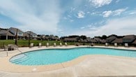 This outdoor community amenity centers around a generously sized freeform pool with a distinctive curved design, filled with clear, blue water that reflects the open sky above. The pool's organic shape adds visual interest and creates a welcoming recreational atmosphere suited for both leisurely swimming and relaxation.

Surrounding the pool is a broad, light-colored concrete sundeck that provides ample space for lounging. Several sling-style lounge chairs are arranged along the poolside, offering comfortable spots to enjoy the outdoors and sunshine. A wrought iron perimeter fence encloses the pool area, providing a clear boundary while maintaining open sightlines to the surrounding landscape.

Beyond the fence, mature trees and well-maintained green lawns frame the space, with a backdrop of residential homes featuring neutral-toned exteriors and pitched rooflines. This neighborhood setting contributes to a sense of community and privacy. The overall ambiance is bright, open, and inviting, with an abundance of natural light and expansive sky views enhancing the outdoor experience throughout the day.