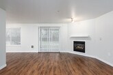 This single-floor space offers a clean, modern aesthetic anchored by warm wood-look flooring that flows continuously through the main living areas. The spacious living room is a standout, centered around a built-in fireplace framed by a white mantel. Natural light fills the room through a large sliding glass door fitted with vertical blinds, opening to a view of mature greenery outside, complemented by an additional side window that further brightens the space.

The kitchen is thoughtfully designed with white shaker-style cabinetry, subway tile backsplash, and gray countertops. Black appliances—including a refrigerator, electric range, and dishwasher—provide a bold contrast against the bright cabinetry. A flush ceiling fixture provides ample overhead lighting, and the layout offers generous counter and storage space.

Two bathrooms serve the unit, each with its own distinct character. One features hexagonal tile flooring, a white vanity with an integrated sink, and a well-lit mirror. The second bathroom includes wood-look flooring, a white vanity with darker countertop, and a glass-enclosed shower stall. Both spaces maintain a clean, neutral palette consistent with the overall design of the unit.