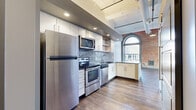 Located at 619 E William St in the heart of downtown Wichita, KS, this one-bedroom, one-bathroom unit blends historic industrial character with contemporary upgrades. The living area is an immediate standout, showcasing dramatic exposed brick walls, soaring ceilings with ornate coffered detailing, and large arched windows that flood the space with natural light and offer views of the surrounding streetscape. Rich, dark hardwood-style flooring runs throughout the main living areas, grounding the open layout with warmth.

The kitchen is well-appointed with stainless steel appliances — including a refrigerator, range, microwave, and dishwasher — complemented by dark granite countertops, white cabinetry, a textured tile backsplash, and open wood shelving. Exposed ductwork and recessed lighting reinforce the loft-inspired aesthetic.

A hallway with tall ceilings and bold dark-paneled doors leads to the bedroom and bathroom. The bedroom features carpeted flooring, a large ornately trimmed window with exposed brick surrounds, and a cozy, well-proportioned layout. The bathroom offers a full soaking tub-shower combination surrounded by earthy glazed stacked tile, wood-look flooring, and clean white fixtures, delivering a refined, spa-like feel within a compact footprint.