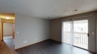This single-floor space presents a clean, functional living area designed with practical simplicity. The room features dark hardwood-style flooring that contrasts with the light gray walls, creating a grounded, neutral aesthetic throughout. A textured popcorn ceiling adds architectural character overhead, while standard HVAC venting ensures climate comfort.

Natural light enters the space through a sliding glass door fitted with vertical blinds, offering both privacy control and access to an adjoining outdoor deck or balcony. The door provides a direct connection to the exterior, enhancing the sense of openness within the room.

The walls are finished in a soft gray tone and accented with warm wood-toned baseboards that complement the flooring. Multiple electrical outlets are distributed along the walls, offering practical functionality. The overall layout is open and unobstructed, providing flexibility for various furniture arrangements. The space maintains a straightforward, move-in-ready character well-suited for everyday living.