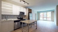 This well-designed two-bedroom, one-bathroom residence offers a thoughtfully laid-out single-level floor plan with contemporary finishes throughout. The open-concept kitchen flows seamlessly into the living area, creating a cohesive and functional space. The kitchen is equipped with stainless steel appliances, including a refrigerator, range, microwave, and dishwasher, complemented by white gloss upper cabinetry with frosted glass accents, warm-toned lower cabinetry, a mosaic tile backsplash, and granite countertops. A portable granite-topped island provides additional prep and dining space. Light hardwood-style flooring runs throughout the main living areas, enhancing the bright and airy feel. The living room benefits from expansive floor-to-ceiling windows that invite abundant natural light and offer views of the surrounding neighborhood. A frosted glass sliding door adds a modern touch and provides flexible room division. Both bedrooms are well-proportioned, with one featuring a spacious closet. The bathroom showcases an elegant herringbone-patterned tile floor, a vessel sink atop a granite vanity with illuminated mirror, and a full tub-shower combination with white subway tile and a contrasting black tile accent border. The overall aesthetic is clean, modern, and move-in ready.