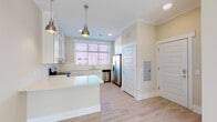 Located at 900 Carraway Xing in Chapel Hill, NC, this single-floor unit offers a thoughtfully designed layout with contemporary finishes throughout. The kitchen serves as a central highlight, featuring crisp white shaker-style cabinetry with glass-front upper cabinets, light quartz countertops, a subway tile backsplash, and a suite of stainless steel appliances including a refrigerator, range, and dishwasher. A peninsula island with pendant lighting provides additional prep space and casual seating, while a large window above the sink floods the area with natural light and neighborhood views. Light-toned hardwood-style flooring runs throughout the main living areas, contributing to a clean and airy aesthetic. The living area features recessed lighting, crown molding, and direct access to an outdoor space via glass-paneled doors, allowing ample sunlight to pour in. The bedroom is carpeted and includes a ceiling fan and generously sized windows with blinds. Two bathrooms round out the unit — one featuring a tub-shower combination with white subway tile surround, and a second with a separate walk-in glass shower, both finished with white vanity cabinetry, light countertops, and tile flooring. The overall palette of warm neutrals and white creates a bright, welcoming atmosphere throughout.