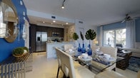 This well-appointed two-story residence offers a thoughtfully designed layout that seamlessly blends form and function across both levels. The open-plan living area serves as the heart of the home, where the kitchen and dining space flow together in a cohesive and inviting arrangement. The kitchen is outfitted with dark espresso cabinetry, stainless steel appliances including a French door refrigerator and built-in microwave, and a clean white peninsula that doubles as a breakfast bar. Recessed lighting and track lighting illuminate the space, complementing the bright, white ceilings and light-toned flooring throughout. The adjacent dining area is spacious enough to accommodate a full-sized glass dining table with seating for multiple guests, elegantly styled with coastal-inspired blue glassware and lush tropical greenery. Large windows allow generous natural light to filter in, enhancing the open and airy ambiance of the living spaces. The overall aesthetic is modern and coastal, with a crisp white and navy blue palette punctuated by natural textures. The two-floor layout provides ample room for comfortable living, making this space as practical as it is visually appealing.