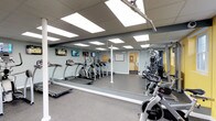 This fitness center is thoughtfully designed to support a well-rounded workout experience. The main exercise area features durable rubber flooring throughout and is illuminated by recessed panel lighting set within a drop-tile ceiling, creating a clean, well-lit environment. A row of treadmills and elliptical trainers lines one side of the space, each positioned in front of individual wall-mounted flat-screen televisions, allowing users to stay entertained during cardio sessions. Full-length mirrors span an entire wall, visually expanding the room while providing functional feedback during exercise. A dedicated strength training area houses a multi-station cable machine, a spin bike, and an additional resistance machine, all situated along a boldly painted yellow accent wall lined with a series of large windows that invite natural light into the space. The windows offer a view of the surrounding outdoor environment, adding an open, airy quality. A framed workout reference chart is displayed on the wall, and a water cooler is accessible through an adjoining passage. The overall ambiance is energetic yet practical, offering residents a comprehensive and inviting fitness environment.
