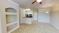 This single-floor residence offers a bright and welcoming open-concept design that seamlessly connects the living area and kitchen. The living space is finished with plush, neutral-toned carpeting and features a charming built-in arched shelving unit along one wall, ideal for display or storage. Crown molding adds a refined architectural touch throughout, complementing the clean, light-colored walls. A ceiling fan is centrally positioned, enhancing both comfort and airflow in the main living area.

The kitchen is defined by a raised peninsula with a dark granite-style countertop, providing both a functional workspace and a casual dining or gathering spot. White cabinetry lines the kitchen walls and upper sections, offering generous storage, while stainless steel appliances — including a refrigerator, microwave, and range — lend a modern, polished look. Recessed and overhead lighting brightens the kitchen efficiently.

Adjacent to the kitchen, a tiled entryway leads to a white paneled front door, creating a clean transition zone between the interior and exterior. The overall ambiance of the space is calm and approachable, with a consistent neutral palette that makes it feel open, airy, and move-in ready.