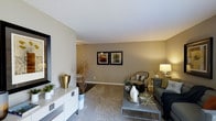 This single-floor residence offers a thoughtfully designed living space that balances modern aesthetics with comfortable functionality. The living area serves as the centerpiece of the home, showcasing neutral warm-toned walls that create a calm and welcoming atmosphere throughout. Soft, plush carpeting underfoot adds warmth to the space, while clean white ceilings enhance the sense of openness.

The living area is well-appointed with contemporary furnishings, including a sleek gray sofa, a metallic-accented coffee table, and a stylish accent chair, all arranged to encourage comfortable conversation and relaxation. A low-profile white media console anchors one side of the room, complemented by decorative accessories including gold vases, greenery, and sculptural arrangements that add personality and depth. Two framed artworks in matching black frames are displayed together on the main wall, providing a cohesive focal point.

Natural light filters into the space, lending a bright and airy quality to the interior. The overall design reflects a cohesive modern sensibility, with careful attention to detail in the layering of textures, metallic accents, and neutral tones. The layout feels spacious and well-proportioned, making it a versatile and elegant environment suitable for both everyday living and entertaining.