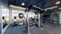 This modern fitness center is designed to support a full range of workout routines in a bright, open environment. The space is anchored by a striking dark-painted exposed ceiling with industrial beams, large ceiling fans, and pendant lighting, lending the gym a contemporary, high-energy aesthetic. Deep navy accent walls contrast with the predominantly open layout and contribute to the space's bold, dynamic character.

Floor-to-ceiling windows and glass-panel garage-style doors flood the interior with natural light and provide sweeping panoramic views of the surrounding landscape and skyline. One wall of large glass doors opens directly onto an outdoor terrace and pool deck, seamlessly blending indoor and outdoor fitness environments.

The gym is comprehensively equipped with a full row of treadmills, elliptical machines, stationary bikes, and rowing equipment positioned to take advantage of the exterior views. A multi-functional training rig serves as the centerpiece of the strength zone, complemented by a dual cable machine, a complete set of kettlebells, medicine balls of varying weights, stability balls, and a neatly organized dumbbell rack. Rubber flooring throughout ensures safety and durability. A wall-mounted mirror extends the visual depth of the space, and mounted flat-screen televisions provide entertainment. Together, these elements create a professional-grade fitness environment suited for cardio, strength, and functional training.