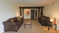This single-floor residence offers a welcoming and functional layout designed for comfortable everyday living. The living room serves as the heart of the space, featuring a pair of rich dark leather sofas arranged symmetrically around a sleek glass-top coffee table, creating an inviting seating area ideal for relaxing or entertaining. A bold patterned area rug anchors the arrangement and adds a touch of visual interest to the soft, neutral carpeted flooring that runs throughout. One of the most distinctive architectural elements is the dark wood exposed beam that spans across the ceiling, adding depth and character to the otherwise light and airy room. The neutral-toned walls provide a versatile backdrop, complemented by warm accent lighting that contributes to a cozy ambiance. Decorative touches, including framed wall art and tall accent plants, add personality to the space. A hallway visible from the living area leads to additional rooms, suggesting a practical and well-connected floor plan. The overall design balances simplicity with warmth, making it a comfortable and livable single-floor home.