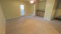 This single-story space offers a comfortable and functional layout highlighted by several notable built-in features. The main living area is finished with wall-to-wall carpet in a neutral tone, complementing the warm beige walls throughout. A ceiling fan with integrated lighting is centrally mounted, providing both air circulation and ambient illumination.

A standout feature of the space is the built-in wooden shelving unit along one wall, offering open display or storage shelving above a lower cabinet section with a dark stone-style countertop. The cabinetry features traditional-style wooden doors and drawers, adding a warm, classic character to the room.

Natural light enters through a glass-panel exterior door fitted with blinds, which also provides access to the outdoors. The door is framed by white trim that contrasts cleanly against the neutral walls. An open doorway visible from the main area suggests additional rooms beyond, contributing to a sense of connected, flowing space. Overall, the space presents a practical and inviting environment with functional built-ins and a cohesive, understated aesthetic.