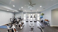 This single-floor fitness center is thoughtfully designed to support a comprehensive workout experience. The space is finished with soft blue-gray walls that create a calm, focused atmosphere, complemented by a dark rubber flooring that is both durable and practical for exercise use. Recessed lighting is evenly distributed throughout the ceiling, ensuring bright, consistent illumination across the entire floor, while two ceiling fans promote air circulation and comfort during workouts.

Natural light flows generously into the space through a set of white-framed French doors along the far wall, adding an airy, open feel to the interior. The cardio section is well-stocked with multiple treadmills and stationary spin bikes equipped with digital monitors, allowing users to track their performance. A dedicated free weights area along one wall includes a rack of exercise balls in varying sizes, kettlebells, dumbbells, and a flat workout bench, catering to strength and flexibility training. Additional equipment such as an elliptical machine further broadens the range of available workouts. The layout is spacious and organized, with clear zones for different types of exercise, making the fitness center both functional and easy to navigate.