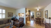 This single-floor residence offers a thoughtfully arranged open-concept layout that seamlessly connects the living, dining, and kitchen areas. The living space is anchored by a patterned upholstered armchair and a plush area rug, complemented by a side table with a lamp and decorative greenery. Framed artwork adorns the walls, adding a touch of character to the room.

A partial dividing wall with a pass-through window separates the living area from the kitchen, which features warm wood cabinetry, light countertops, and white appliances, including a refrigerator. The kitchen layout makes efficient use of space while maintaining an open connection to the adjoining areas.

The dining area, visible to the right, accommodates a wooden table and chairs beneath a ceiling fan with a light fixture, providing both functionality and ambiance. Light hardwood flooring flows throughout the main areas, contributing to a cohesive, warm aesthetic.

A hallway visible beyond the kitchen leads to a bedroom, suggesting a well-organized floor plan. The overall ambiance is cozy and inviting, with consistent warm tones, ample artificial lighting, and a practical yet comfortable design throughout.