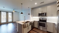 This single-floor space showcases a thoughtfully designed open layout with distinct living zones connected by rich, dark hardwood-style flooring. The kitchen serves as a focal point, featuring gray flat-front cabinetry, a white subway tile backsplash, and a full suite of stainless steel appliances including an electric range, over-the-range microwave, refrigerator, and dishwasher. A peninsula with a white quartz countertop extends into the living area, housing a deep stainless steel farmhouse sink and framed by two industrial-style pendant lights, creating an ideal space for casual dining or entertaining. A built-in wine rack adds a refined touch to the kitchen's design.

The bathroom is clean and contemporary, finished with large-format tile flooring, a walk-in glass-enclosed shower with tile surround, a white undermount sink vanity, and direct access to a well-organized walk-in closet with built-in shelving.

The bedroom is carpeted for comfort and features a ceiling fan and a window offering views of greenery, filling the room with natural light. The overall ambiance is modern and cohesive, with a neutral palette throughout.
