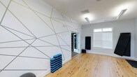 The yoga room is a thoughtfully designed single-floor studio space built to support mindful movement and focused practice. The most striking feature is the large-scale geometric mural adorning the primary wall, composed of intersecting angular lines in gold and gray tones against a crisp white background, creating a dynamic and energizing visual backdrop. The warm, natural hardwood floors run throughout the room, providing a clean and grounded surface ideal for yoga practice. Vaulted ceilings add a sense of openness and airiness to the space, enhancing the overall feeling of calm and spaciousness. Natural light filters in through a set of windows fitted with white blinds, offering brightness while maintaining privacy and glare control. Overhead lighting provides consistent illumination for sessions at any time of day. A rolled black yoga mat is propped along one wall alongside a stacked set of blue foam yoga blocks, indicating the room is well-equipped for various practice styles. A doorway connects the studio to an adjacent storage or equipment area, ensuring props and accessories remain organized and accessible. The overall ambiance is modern, clean, and purposefully minimalist.