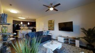 This single-floor residence showcases an open-concept floor plan that integrates the living area and kitchen into one cohesive, functional space. The living room is anchored by a dark navy sectional sofa accented with blue throw pillows, positioned around a white rectangular coffee table atop a patterned area rug. A large flat-screen television is wall-mounted above a contemporary media console, complemented by a decorative metallic wall art piece that adds a bold visual accent. A ceiling fan with a built-in light fixture is centrally positioned overhead, contributing to both airflow and ambient lighting. The kitchen, visible from the living area, features dark cabinetry, pendant lighting over a breakfast bar with bar seating, and modern appliances. The open sightlines between the kitchen and living space create an inviting atmosphere well-suited for entertaining. Light-toned walls throughout enhance the brightness of the interior, while wood-style flooring adds warmth and continuity across the space. Greenery in the foreground introduces a natural element that softens the modern aesthetic.