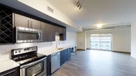 This single-floor space showcases a sleek, modern design with an open-concept layout that seamlessly connects the kitchen and living area. The kitchen is well-appointed with dark espresso cabinetry contrasted by crisp white quartz countertops and a classic white subway tile backsplash. Stainless steel appliances — including a range, built-in microwave, and dishwasher — add a polished, professional touch. A deep undermount sink is centrally positioned along the countertop, complemented by a contemporary brushed-nickel faucet. Track lighting overhead provides focused illumination throughout the kitchen space. The flooring transitions smoothly from the kitchen into the open living area, finished in light-toned hardwood-style planks that enhance the airy feel of the space. Exposed ductwork along the ceiling adds an industrial character to the living area. Large floor-to-ceiling windows flood the living space with natural light and offer views of the surrounding urban environment. The overall ambiance is clean, contemporary, and functional, making it an inviting and practical space for everyday living.