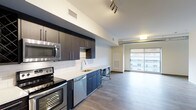 This contemporary apartment showcases a thoughtfully designed open-concept layout, seamlessly blending the kitchen and living areas. The kitchen is a standout feature, boasting dark wood cabinetry that contrasts beautifully with stainless steel appliances, including a built-in microwave and oven. A subway tile backsplash adds a touch of classic elegance, while the expansive countertop provides ample workspace. The room is illuminated by both recessed lighting and a modern track light fixture, enhancing the space's modern aesthetic. Large windows flood the area with natural light and offer city views, creating an airy atmosphere. The hardwood flooring extends throughout, unifying the space and adding warmth. A vaulted ceiling with exposed ductwork lends an industrial chic vibe, while also making the room feel more spacious. This apartment is ideal for those who appreciate contemporary design and functional living spaces.