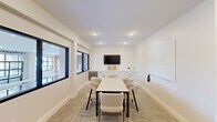 Located at 3688 Brinell St W in Columbus, Ohio, this compact yet well-appointed conference room offers a clean, modern design suited for focused meetings and collaborative work. The single-floor space is finished with crisp white walls and recessed ceiling lighting that creates a bright, professional atmosphere throughout.

At the center of the room sits a rectangular white meeting table surrounded by upholstered, curved chairs in a neutral tone, comfortably accommodating a small group. Along one wall, a row of white cabinetry with dark hardware provides ample storage, topped with a quartz-style surface and complemented by a built-in beverage refrigerator. A wall-mounted flat-screen TV is positioned above the cabinetry, making the space well-equipped for presentations.

Interior windows with bold black frames connect the room visually to an adjacent common area, allowing natural light to filter in while maintaining a sense of openness. A large whiteboard mounted on the wall supports brainstorming and collaborative sessions. A wall-mounted split-system air conditioning unit ensures independent climate control.

Patterned, textured carpet in warm neutral tones lines the floor, adding comfort underfoot while contributing to the room's polished aesthetic. A dedicated exit door with a keypad entry system provides secure, private access to this functional and stylish meeting space.