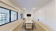 Located at 3688 Brinell St W in Columbus, Ohio, this compact yet well-appointed conference room offers a clean, modern design suited for focused meetings and collaborative work. The single-floor space is finished with crisp white walls and recessed ceiling lighting that creates a bright, professional atmosphere throughout.

At the center of the room sits a rectangular white meeting table surrounded by upholstered, curved chairs in a neutral tone, comfortably accommodating a small group. Along one wall, a row of white cabinetry with dark hardware provides ample storage, topped with a quartz-style surface and complemented by a built-in beverage refrigerator. A wall-mounted flat-screen TV is positioned above the cabinetry, making the space well-equipped for presentations.

Interior windows with bold black frames connect the room visually to an adjacent common area, allowing natural light to filter in while maintaining a sense of openness. A large whiteboard mounted on the wall supports brainstorming and collaborative sessions. A wall-mounted split-system air conditioning unit ensures independent climate control.

Patterned, textured carpet in warm neutral tones lines the floor, adding comfort underfoot while contributing to the room's polished aesthetic. A dedicated exit door with a keypad entry system provides secure, private access to this functional and stylish meeting space.