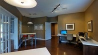 This single-floor space offers an open, multifunctional layout that combines living, dining, and work areas within one cohesive environment. Dark hardwood flooring runs throughout, providing a warm and grounded aesthetic that contrasts with the earthy, olive-toned walls. The space is illuminated by a combination of a flush overhead light fixture and a track lighting system with adjustable heads, allowing for flexible and targeted lighting across the room.

A compact bar-height dining table with two red stools serves as an informal eating or gathering area, adding a pop of color to the otherwise neutral palette. Adjacent to this, a dedicated workspace features a built-in or wall-mounted desk surface with overhead shelving, accommodating a desktop computer and monitor — a practical setup for productivity. Two framed pieces of wall art add a decorative touch nearby.

A doorway leads to a separate room or hallway, suggesting additional private space beyond the main open area. The overall ambiance is contemporary and efficient, with thoughtful use of every area to balance comfort, functionality, and style in a compact yet well-organized setting.